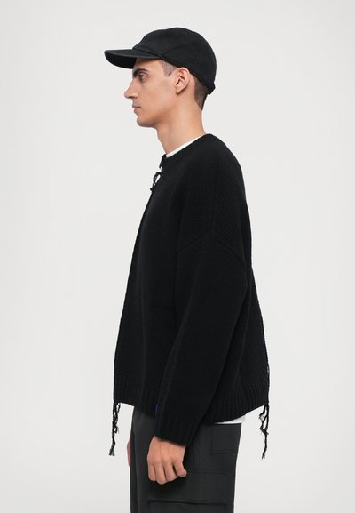Black knitted sweater with a loose fit, round neckline, and fringe detailing at the hem. Paired with a black cap and layered over a white shirt.