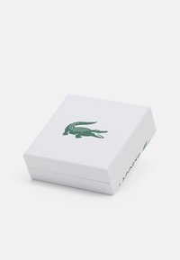 Caja blanca texturizada con el logo de cocodrilo verde y el texto "LACOSTE" en el lateral. Forma cuadrada, acabado suave, diseño minimalista.