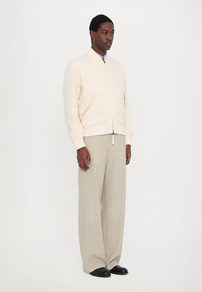 Veste bomber beige clair avec fermeture éclair, poignets côtelés et deux poches avant, associée à un pantalon large gris clair et des chaussures noires.