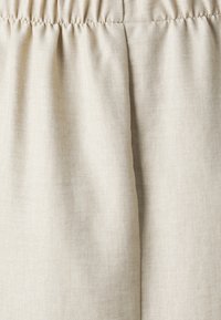 Lys beige stof med en let tekstur, der har en samlet elastik i taljen. Glat overflade med subtil glans og naturlig fald.