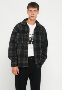 Jachetă în carouri, navy și bej din fleece, având guler și guler de tip colier, buzunare frontale și închidere cu nasturi, asociată cu un tricou alb cu grafică.