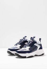 Marinblå och vita sportiga sneakers med nät- och mockamaterial, utrustade med en kraftig sula, snören och en logotyp på sidan.