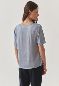 Blouse légère bleu clair à manches courtes avec un col rond, texture lisse et fentes latérales, présentée sur un modèle vu de dos.