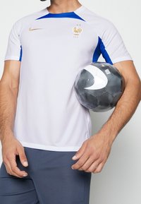Camiseta de fútbol blanca de Nike con acentos azules y emblema dorado, combinada con pantalones cortos deportivos gris oscuro; sosteniendo un balón de fútbol blanco y negro.