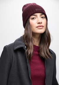 Gorro de punto burdeos con textura acanalada y una etiqueta, combinado con un abrigo oscuro y un suéter burdeos, creando un look acogedor y estratificado.