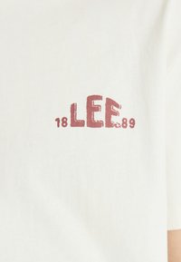 Weißes Baumwoll-T-Shirt mit einem verblassten, burgunderfarbenen Grafikdruck, auf dem "LEE 1889" steht. Kurzärmelig und mit Rundhalsausschnitt. Glatte Textur.