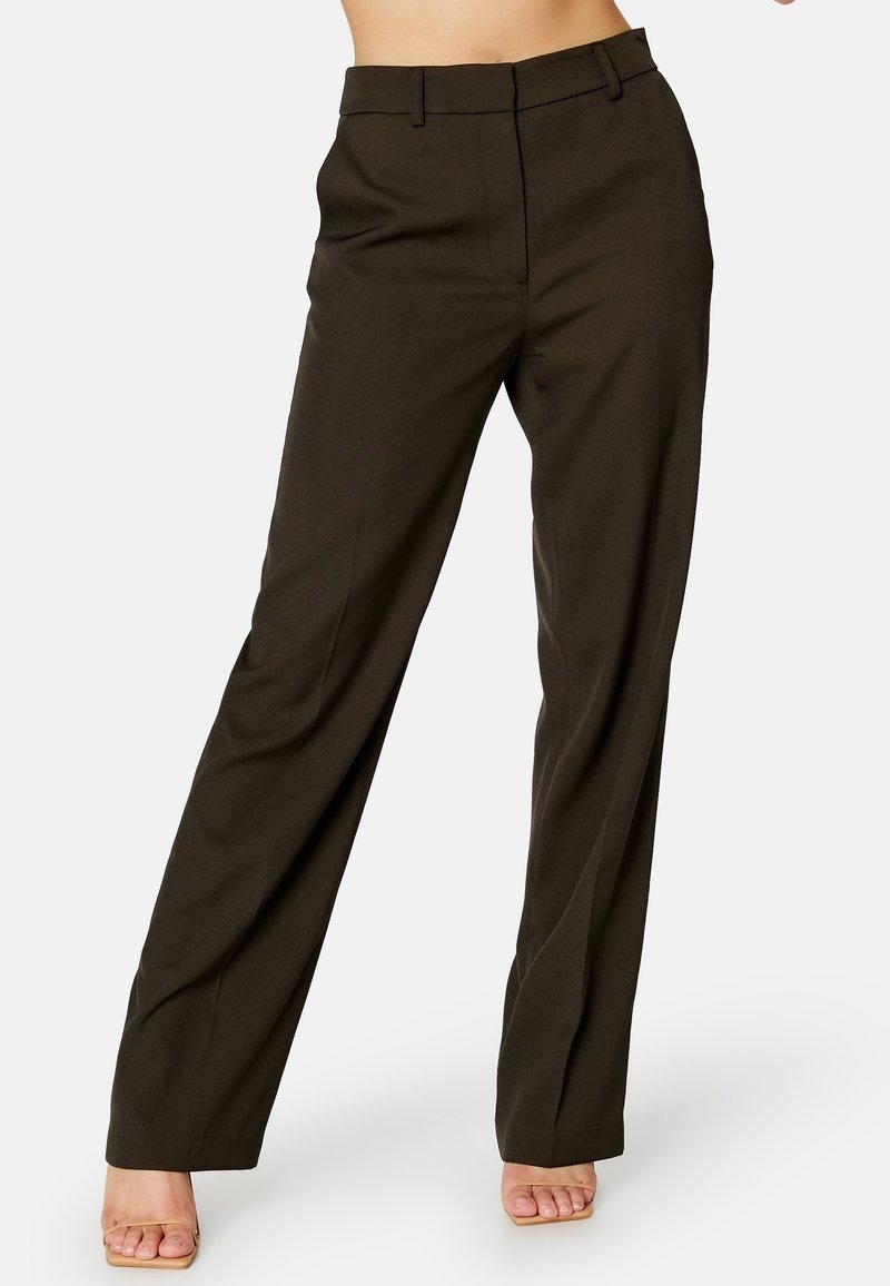 Femme portant un pantalon large taille haute marron foncé associé à des talons beiges à bout ouvert, debout devant un fond blanc uni.