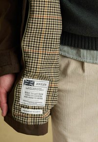 Brązowa kurtka z włożonym w creamową, czarną i czerwoną kratkę; posiada metkę z informacjami o marce i instrukcjami dotyczącymi pielęgnacji.