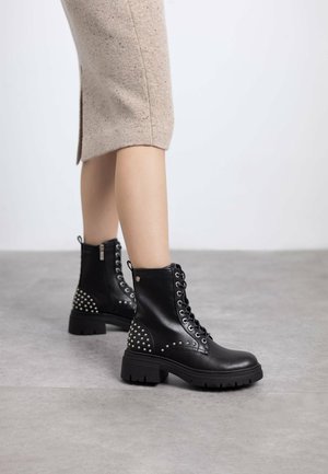 Botas de tobillo de cuero negro con diseño de cordones, detalles con tachuelas en el talón y una suela de goma gruesa para mayor tracción.