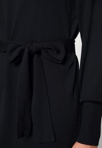 Vêtement en tricot noir avec un détail de ceinture nouée à la taille, présentant une surface texturée et des poignets de manches côtelés.