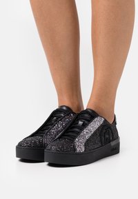 Czarne buty sportowe z błyszczącego materiału, z szarym błyszczącym paskiem, teksturowaną powierzchnią i grubą czarną podeszwą.