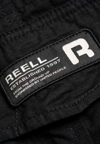 Schwarzes Stoffetikett mit "REELL" in fetten weißen Buchstaben, gegründet 1997, mit zusätzlichem Text darunter in kleinerer Schrift. Strukturierter Hintergrund.