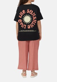 Schwarzes Baumwoll-T-Shirt mit einem Sonnenmotiv in Creme und Pink auf der Rückseite. Lockerer Schnitt, kombiniert mit rosa weit geschnittenen Hosen und Sandalen.