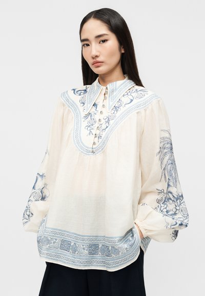 Γυναίκα που φοράει μια άνετη, εκρού μπλούζα με μπλε ανθιστή κεντήματα στο γιακά, τα μανίκια και το τελείωμα, συνδυασμένη με σκούρο παντελόνι, στέκεται μπροστά σε λευκό φόντο.