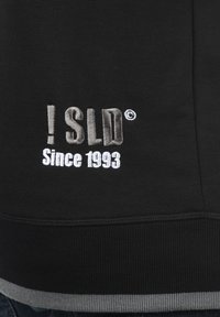 Zwarte sweatshirt met geborduurde tekst "! SLD" in grijs en "Sinds 1993" in wit, met een geribbelde grijze zoom en zachte textuur.