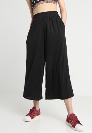 Personne portant un pantalon noir large et court et des baskets rouges, debout devant un fond clair uni.