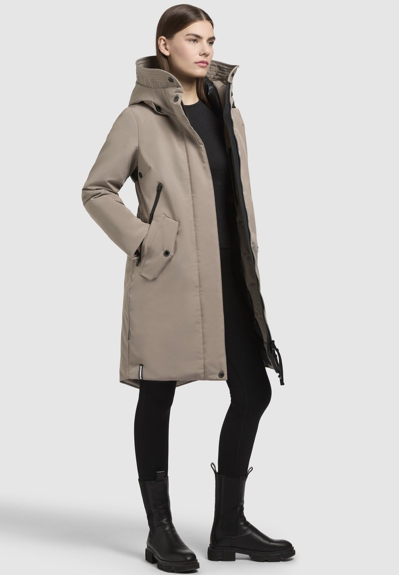 Parka color khaki con colletto alto, zip davanti, tasche laterali e cordini regolabili, abbinata a pantaloni neri e stivali.