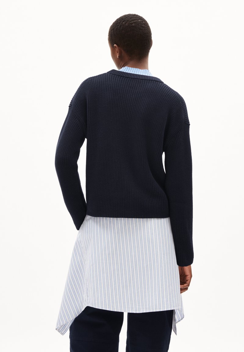 Marineblauer Strickpullover mit cropped Design, langen Ärmeln und gerippter Textur, über einem blau-weiß gestreiften Hemd geschichtet.