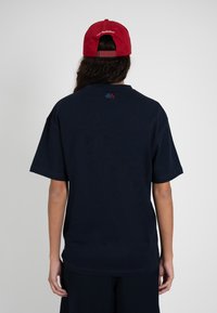 Persona con cabello largo y oscuro que lleva una gorra roja hacia atrás y una camiseta azul marino lisa, mirando de espaldas a la cámara contra un fondo neutro.