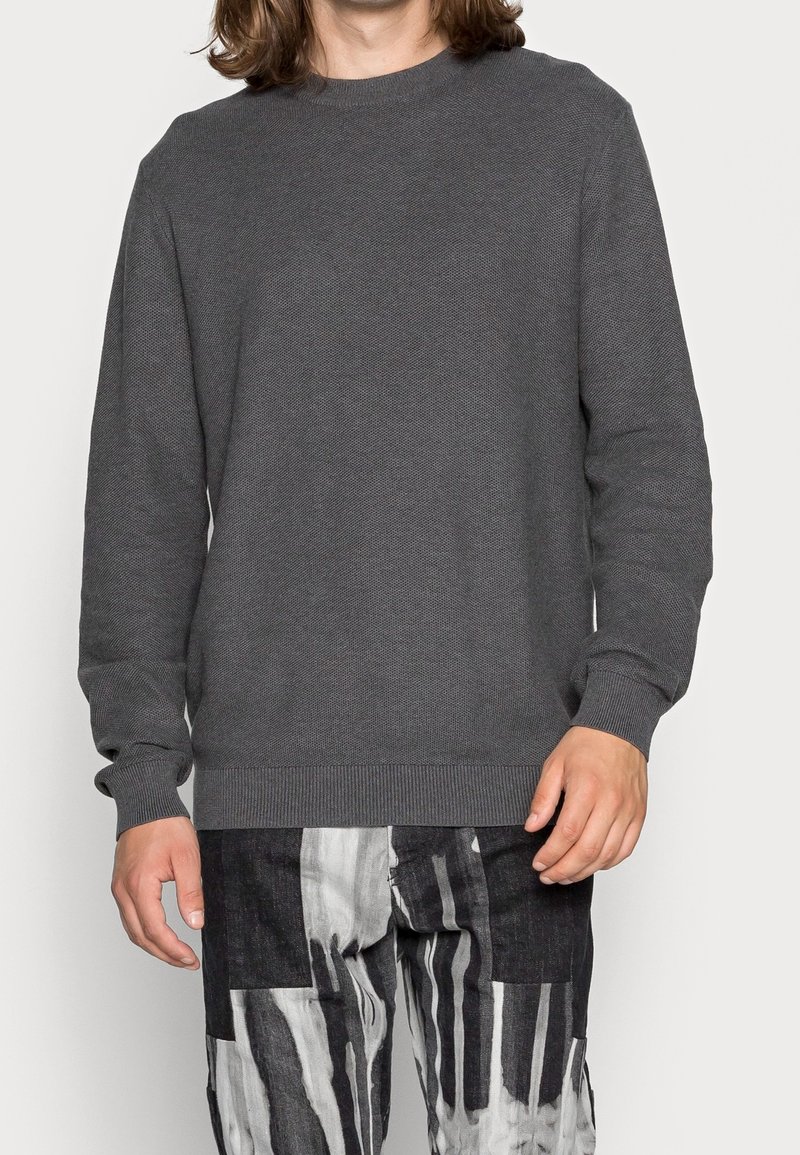 Personne portant un pull texturé gris foncé à manches longues et un pantalon à rayures verticales noir et blanc, visible du cou jusqu'aux hanches.
