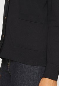 Černý pletený cardigan s knoflíkovým zapínáním, žebrovanými manžetami a náprsní kapsou. Má měkkou texturu a přiléhavý střih.