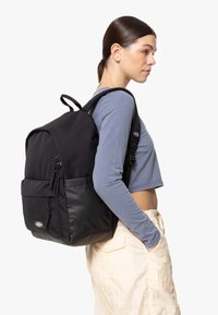 Zaino in tessuto nero con esterno texturizzato e fondo in pelle lucida, dotato di doppie tasche frontali con zip e spallacci regolabili.