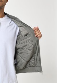 Giacca trapuntata grigio chiaro con chiusura a zip, polsini a coste e foderatura interna liscia, che presenta tasche interne e un fit rilassato.