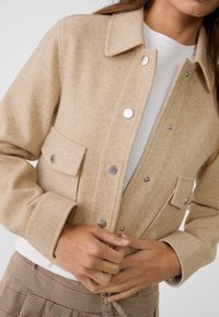 Beige směsová vlnaový kabát s límečkem, stříbrnými druky a dvěma předními kapsami. Měkká textura, volný střih a strukturovaný design.