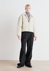 Veste matelassée crème avec fermeture éclair sur le devant, surface texturée et design cropped, assortie à un pantalon large noir et un col roulé rayé.