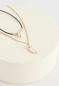 Schichtkette mit einer goldenen Kette, einem weißen Anhänger, einem silbernen Tropfenakzent und einem schwarzen Band mit goldenen Perlen.