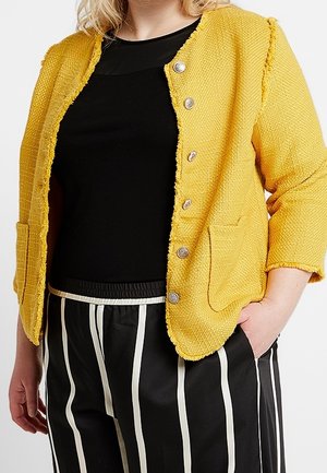 Person iført en tekstureret gul knapjakke over en sort top og sorte bukser med lodrette hvide striber, den ene hånd i lommen.