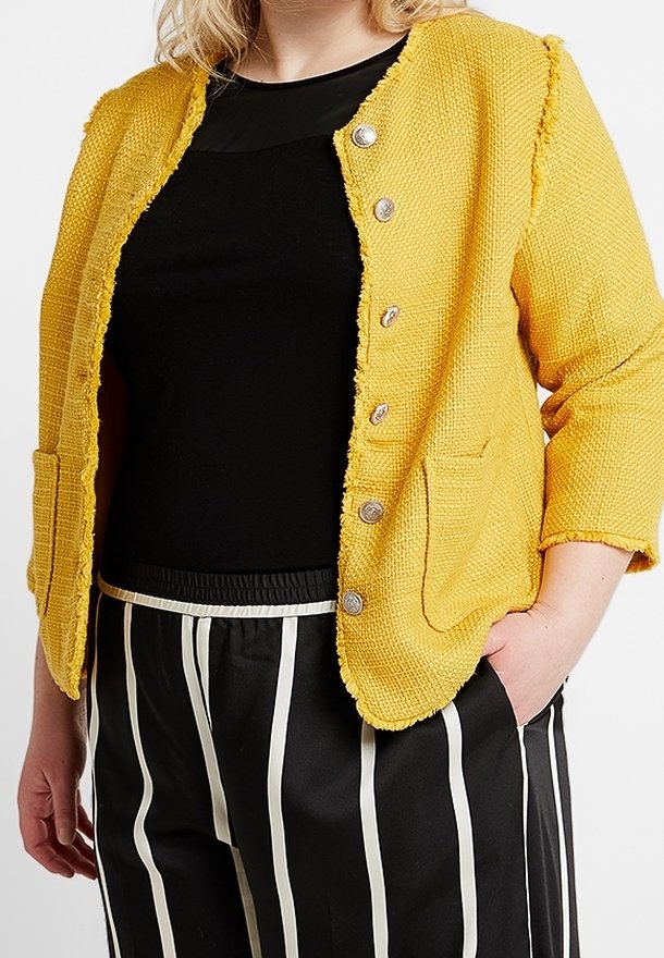 Personne portant une veste jaune texturée boutonnée par-dessus un haut noir et un pantalon noir à rayures blanches verticales, une main dans la poche.