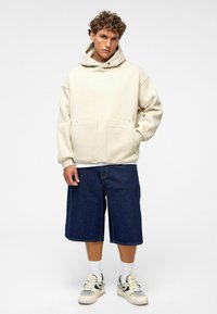 Beige oversized hoodie met een kangoeroezak, gedragen met donkerblauwe denim shorts. Witte sneakers met zwarte accenten, gecombineerd met witte sokken.