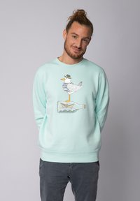 Un sweat en turquoise présente un canard de dessin animé portant une chemise rayée et un chapeau, se tenant sur une bouteille avec un bateau en papier à l'intérieur.