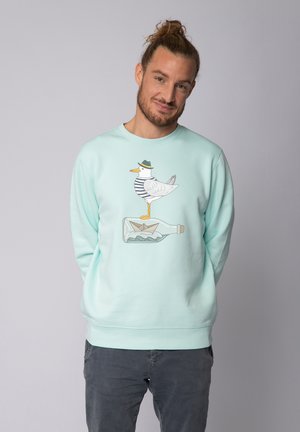Turquoise sweatshirt met een cartoon-eend die een gestreept shirt en hoed draagt, staande op een fles met een papieren bootje erin.