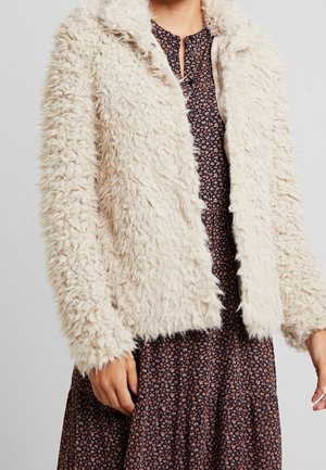 Femme portant une veste duveteuse couleur crème par-dessus une robe florale foncée avec de petites fleurs roses, debout devant un fond uni.