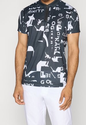 Man die een zwart poloshirt draagt met een witte abstracte kangoeroe en tekstprint, gecombineerd met witte broek, tegen een effen lichte achtergrond.