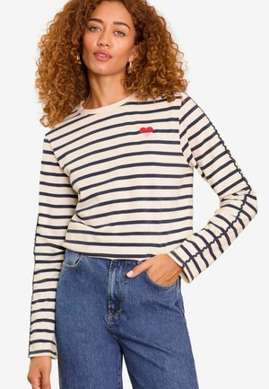 Stribet langærmet top i creme og marineblå med en rød hjerte-detalje. Har ribbede manchetter og et cropped design. Denim jeans synlige.