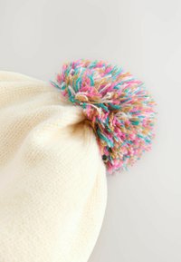 Strikket cremefarvet hue med en flerfarvet pom-pom øverst. Pom-pommen har rosa, blå og brune tråde, som tilføjer tekstur og farvekontrol.