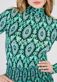 Haut à col roulé à manches longues en vert foncé avec des motifs floraux turquoise et violets Intriqués ; tissu texturé avec un détail de taille froncée.