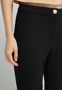 Close-up van een persoon die zwarte broek draagt met een gouden knop en een beige top, met de hand die aan de zijkant rust.