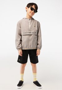 Vaaleanharmaa huppari, jossa musta logo ja yksityiskohdat, mustat shortsit, kelta-raita sukat ja mustat lenkkarit. Rauhallinen ilme, aurinkolasit päässä.