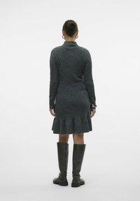 Abito grigio in maglia con texture a coste, maniche lunghe e orlo svasato. Abbinato a stivali in pelle verde alto fino al ginocchio. Vista da dietro.