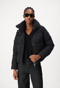 Black quilted puffer jacket with a high collar, featuring a zip closure and snap buttons. Worn over a black V-neck top and dark pants.