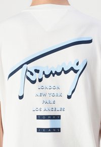 Bijela pamučna majica s plavim grafičkim dizajnom koji sadrži stilizirani tekst "Tommy" i nazive gradova: London, New York, Pariz, Los Angeles.