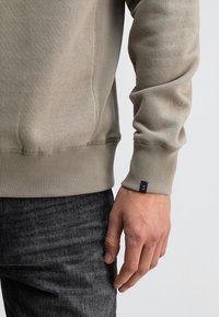 Lichtgrijze sweatshirt met ribgebreide manchetten en onderrand, met een klein zwart logotag op de manchette. Draag het over donkergrijze jeans.