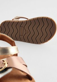Sandalo in pelle marrone con cinturino metallico oro e argento, dotato di una suola in gomma spessa e ondulata con dettagli brandizzati e un accento con fibbia.