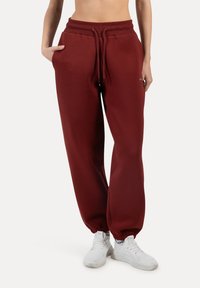 Burgunderfarbene Sweatpants aus weichem Material mit einem Kordelzugbund, schmal zulaufenden Abschlüssen und Seitentaschen. Kombiniert mit weißen Sportschuhen.