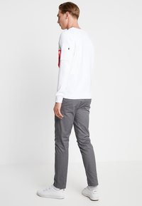 Camicia bianca a maniche lunghe con accento rosso sulle maniche, abbinata a pantaloni aderenti grigi. Sneakers bianche completano il look. Texture del tessuto lisce visibili.