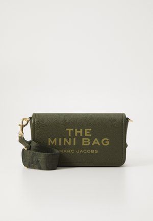 Olijfgroene Marc Jacobs mini tas met gouden letters en een afneembare bijpassende schouderriem met gouden hardware op een witte achtergrond.
